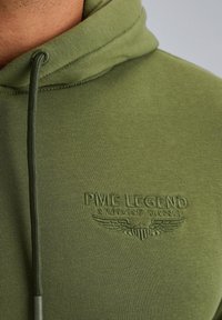 Felpa color verde oliva con una texture morbida. Presenta un cappuccio con cordino e un logo in rilievo "PME LEGEND AMERICAN CLASSIC" con dettagli a forma di ala.