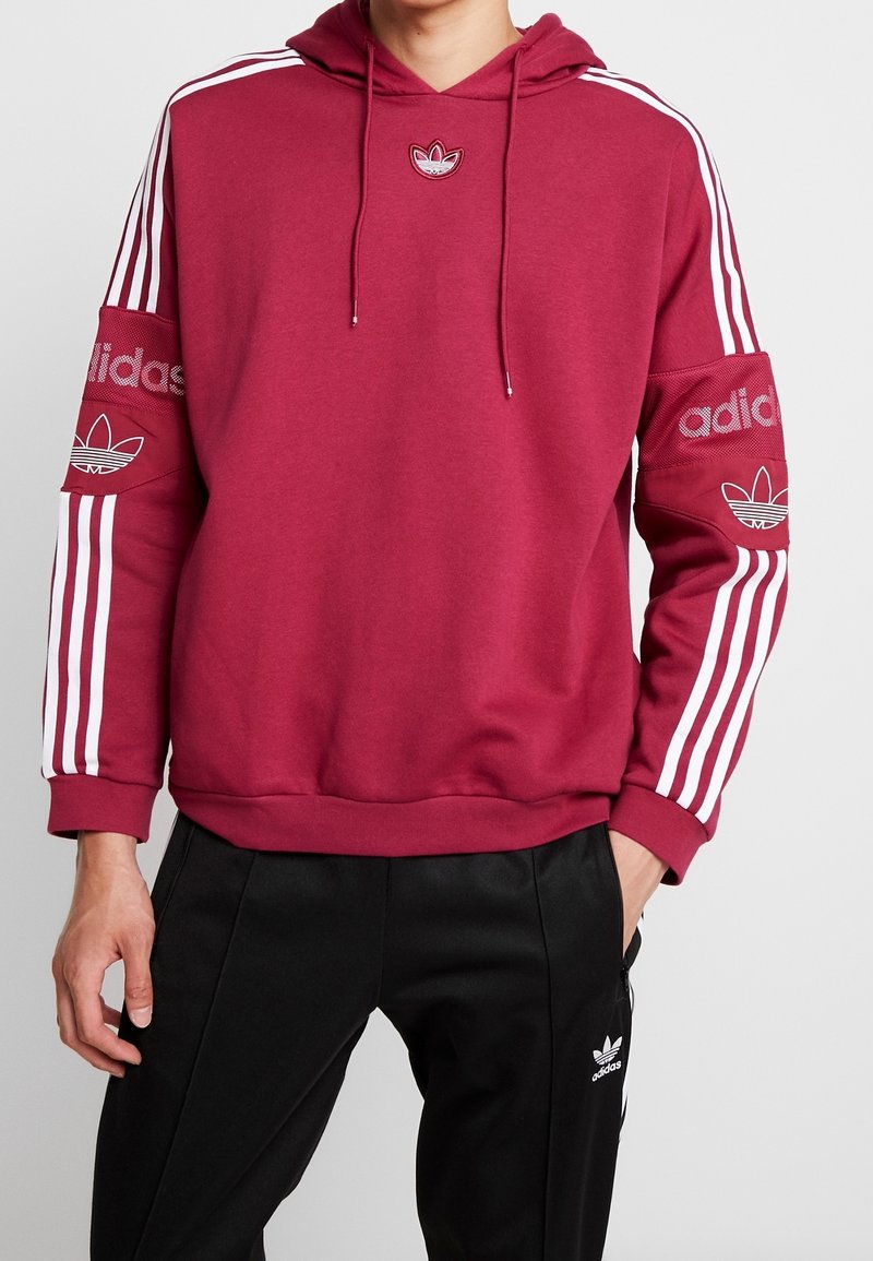 Vinröd huvtröja med vita ränder på ärmarna, prydd med broderade Adidas-logotyper på bröstet och ärmarna, tillverkad av mjukt tyg.