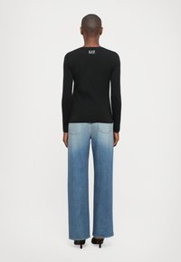 Persoon in een zwart shirt met lange mouwen met het "EA7" logo op de bovenrug, vervaagde wijd gesneden blauwe jeans en zwarte hakken, staand met de rug naar de kijker gericht.