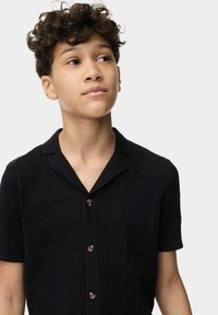 Camisa negra de manga corta con un patrón texturizado, cuello con muesca y cierre de botones. La tela parece suave y tiene una textura lisa.