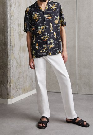 Homme portant une chemise noire à manches courtes imprimée hawaïenne, un pantalon blanc à jambes droites et des sandales noires, debout contre des murs en béton.