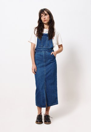 Robe salopette en denim à coupe droite, dotée d'une fente à l'avant, de bretelles ajustables et de deux poches latérales, portée avec des chaussures à lacets noires.