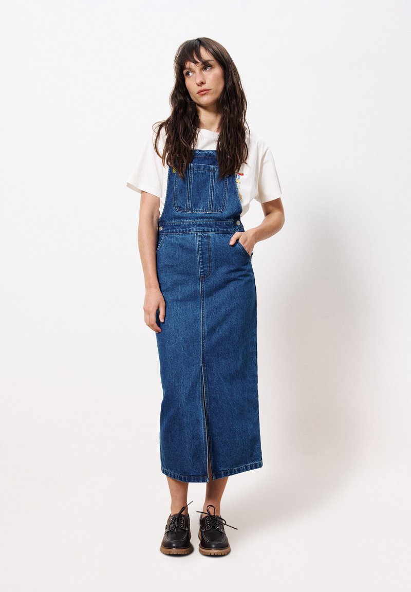 Robe salopette en denim à coupe droite, dotée d'une fente à l'avant, de bretelles ajustables et de deux poches latérales, portée avec des chaussures à lacets noires.