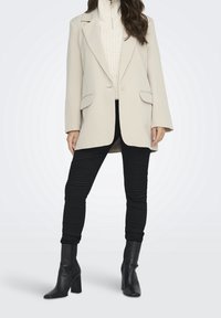 Blazer beige claro con corte oversized, dos bolsillos frontales, mangas largas. Lleva puesto un suéter de punto crema y leggings negros texturizados con canalé.