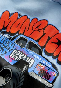 Grafikus póló kék monstrum teherautó mintával, merész piros betűkkel, amelyek a "MONSTER" szót írják, és különböző fekete és rózsaszín graffitivel díszítve.