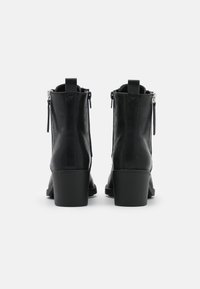 Botas de cuero negro hasta el tobillo con un tacón grueso, cuentan con cremalleras laterales y un acabado suave. En la parte trasera incluyen un lazo para facilitar su colocación.