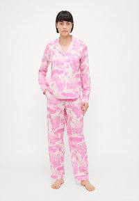 Pink and white patterned pajama set featuring palm trees and animals, with a button-up top and relaxed-fit pants. Soft fabric texture.