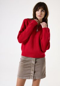 Pull en tricot rouge à côtes avec un col zippé, associé à une jupe en velours côtelé beige avec des détails de boutons à l'avant.
