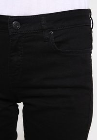 Mustat farkut, jotka on muotoiltu istuviksi, joissa on etutaskut ja metallinen nappikiinnitys vyötäröllä, ommelluin yksityiskohdin.