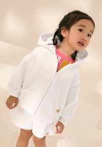 Felpa bianca con zip e vestibilità oversize, tasche a marsupio e polsini a costine, abbinata a pantaloni corti bianchi coordinati. Camicia colorata e texturizzata sotto.