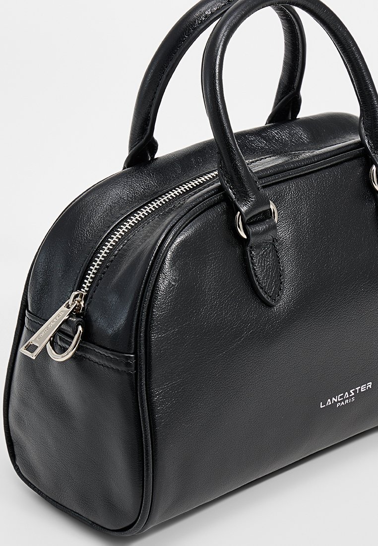 Schwarze Lederhandtasche in abgerundeter Form, mit doppelten Griffen, silbernem Reißverschluss und geprägtem "LANCASTER PARIS"-Logo auf der Vorderseite. Glatte Textur.