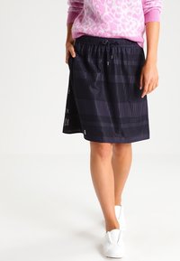 Jupe bleu marine avec une taille élastique, présentant un motif tissé texturé et un design à longueur de genoux. Portée avec des chaussures blanches.