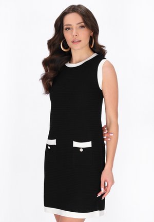 Hosszú barna hajú nő ujjatlan fekete ruhában, fehér szegéllyel és elülső zsebekkel, fehér háttér előtt pózol.