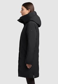 Černý dlouhý péřový kabát s vysokým límcem a kapucí. Má zapínání na zip, boční kapsy a zaoblený spodní okraj, vyrobený z prošívaného voděodolného materiálu.