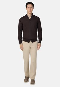 Temno rjava zadrgasta jopica z dvignjenim ovratnikom, rebrastimi manšetami in robom; v kombinaciji s svetlo bež hlačami in rjavimi čevlji.