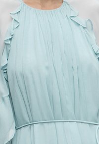 Robe plissée bleu clair avec manches courtes à volants et fine ceinture assortie à la taille, portée par une personne.