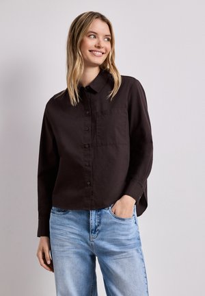 Mujer sonriendo y mirando de lado, vestida con una camisa negra abotonada con bolsillo en el pecho y vaqueros azul claro con una mano en el bolsillo.