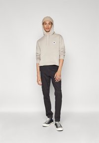 Beige hettegenser med frontlomme og logo-patch, kombinert med svarte cargo-bukser og svart-hvite joggesko. Modellen står mot en enkel bakgrunn.