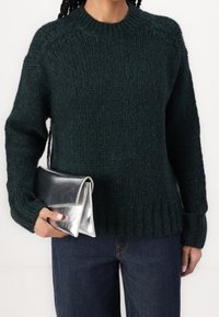 Dunkelgrüner, gestrickter Pullover mit einem gerippten Saum und Raglanärmeln, kombiniert mit einer gefalteten metallisch silbernen Clutch und dunklen Jeans.