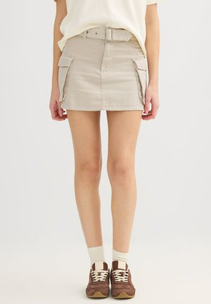 Mujer con mini falda beige con bolsillos laterales, cinturón blanco, calcetines blancos y zapatillas marrones, de pie contra un fondo blanco liso.