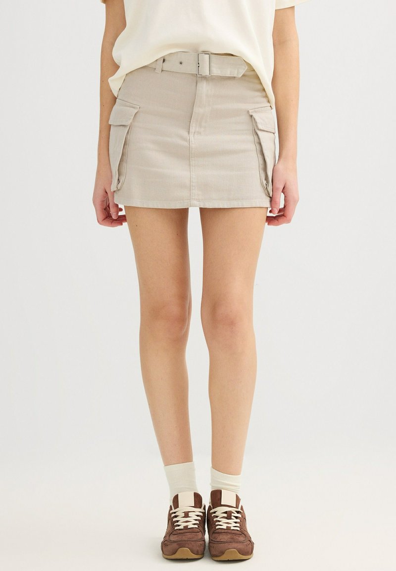 Mujer con mini falda beige con bolsillos laterales, cinturón blanco, calcetines blancos y zapatillas marrones, de pie contra un fondo blanco liso.