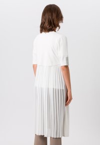 Weißes, plissiertes Kleid mit kurzen Ärmeln, das oben eng anliegt und ab der Taille aus fließendem, durchsichtigem Stoff besteht. Texturierte Akzente an den Ärmeln.