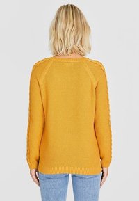 Pull jaune en tricot avec une finition texturée, poignets et ourlet côtelés, et tresses décoratives sur les côtés. Vue de dos.