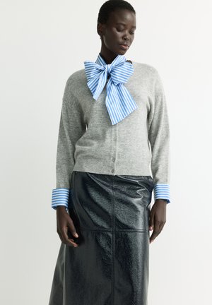 Frau trägt eine graue Strickjacke mit blau gestreiften Bündchen und einer großen blau gestreiften Schleife am Hals, kombiniert mit einem schwarzen Lederrock.