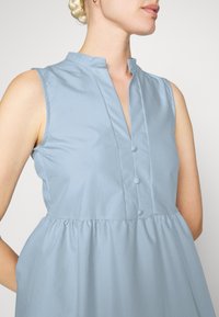 Vestido sem mangas azul claro com gola mandarin, pala de botões e cintura franzida. Tecido suave com acabamento mate.