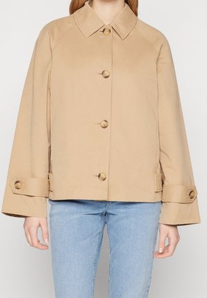 Woman wearing a beige buttoned jacket with wide sleeves and straps, paired with light blue denim jeans.