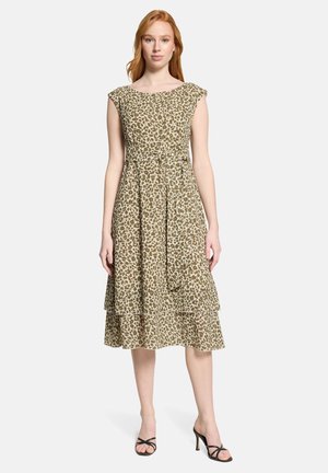 Frau trägt ein ärmelloses, knie-/knielanges Kleid mit Muster und Bindegürtel, steht mit entspannten Armen und schwarzen Sandalen mit Absatz.