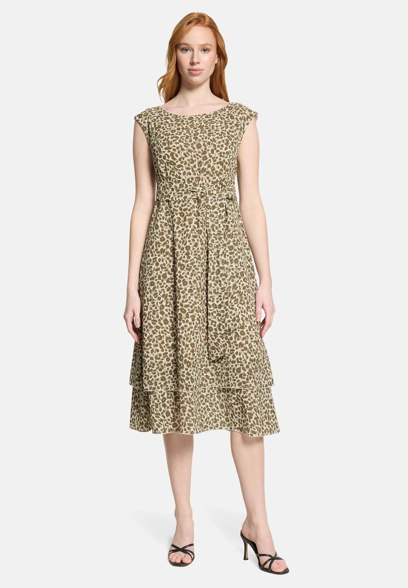 Femme portant une robe sans manches, à motifs, longueur genou, avec une ceinture à nouer, debout les bras détendus, chaussée de sandales noires à talons.