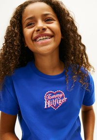 Camiseta de algodón azul con cuello redondo y mangas cortas, que presenta un logo bordado de corazón rojo con "Tommy Hilfiger" en blanco y rosa.