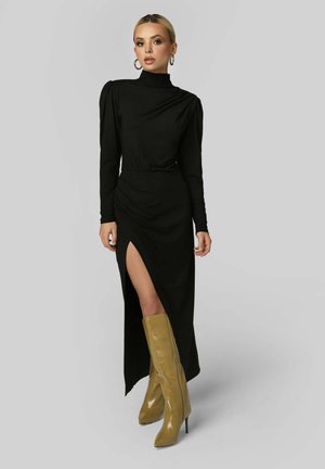 Femme debout vêtue d'une robe noire à manches longues avec une fente haute sur la cuisse et des bottes à talons montantes couleur sable, sur un fond uni.