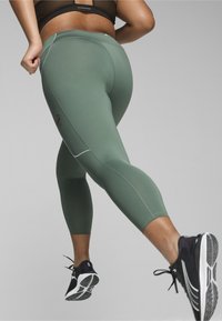 Leggings in un morbido tessuto verde con un design aderente, lunghezza a metà polpaccio e dettagli riflettenti. Indossati con scarpe sportive nere.