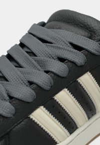 Juodas odinis sportbačio artimas vaizdas su pilkais raišteliais ir trimis tekstūruotomis baltomis šoninėmis juostelėmis virš baltos pado.