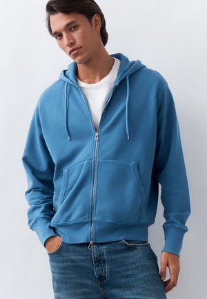 Jeune homme portant un sweat à capuche bleu zippé sur un t-shirt blanc, les mains dans les poches, sur un fond clair uni.