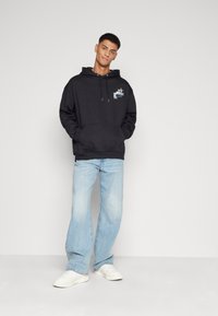 Hoodie noir avec une poche frontale, capuche à cordon de serrage et design graphique sur la poitrine. Porté avec un jean bleu clair et des baskets blanches.