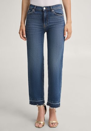 Person trägt gerade geschnittene blaue Jeans mit ausgefransten Säumen, ein weißes Oberteil und goldene offene High Heels und steht vor einem schlichten Hintergrund.
