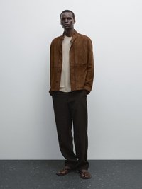Veste en daim marron avec des détails en bouton sur un pull beige clair, associée à un pantalon noir et des tongs marron. Texture lisse et coupe décontractée.