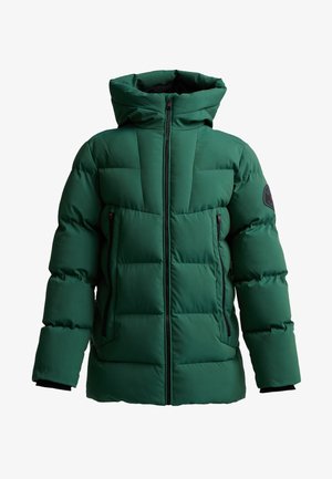 Tienerjongen staand, gekleed in een groene pufferjas met capuchon, zwarte broek en zwarte sneakers tegen een effen lichte achtergrond.