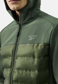 Zielona kurtka Reebok z wzmocnieniem dolnej części. Posiada gładki materiał górny, zamek błyskawiczny i kaptur. Srebrne logo na piersi.