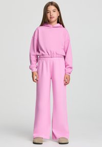 Felpa con cappuccio rosa dal taglio rilassato e vita elasticizzata, abbinata a pantaloni della tuta rosa a gamba larga che mostrano una morbida texture di cotone.