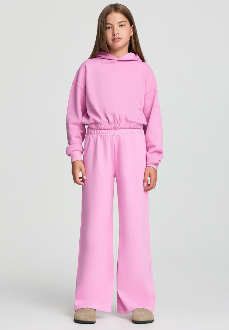 Felpa con cappuccio rosa dal taglio rilassato e vita elasticizzata, abbinata a pantaloni della tuta rosa a gamba larga che mostrano una morbida texture di cotone.