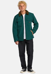 Giacca esterna color teal con collare in pelliccia marrone, due tasche sul petto e chiusura a zip. Indossata con pantaloni neri e sneakers. Materiale in cotone.