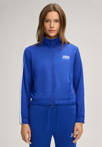 Giubbotto blu con zip, realizzato in tessuto liscio, con strisce bianche a contrasto sulle maniche e un logo sul petto. Design con cintura regolabile.