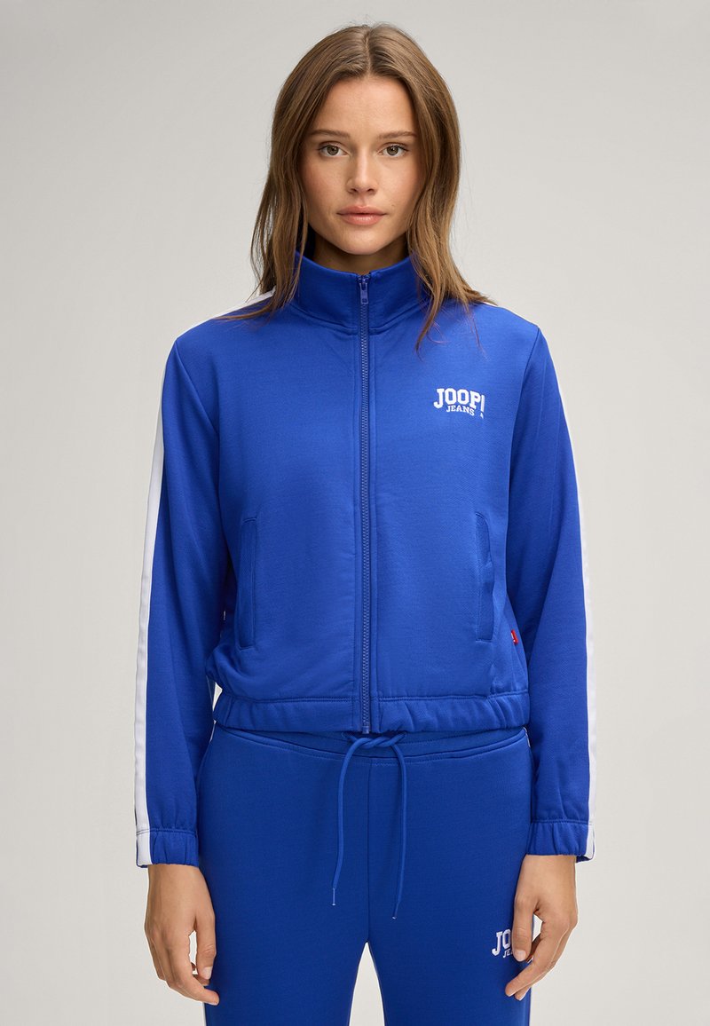 Giubbotto blu con zip, realizzato in tessuto liscio, con strisce bianche a contrasto sulle maniche e un logo sul petto. Design con cintura regolabile.