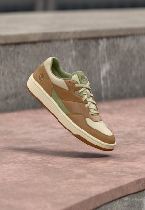 Sneaker with a cream upper, tan leather accents, green details, and a brown rubber sole. Features laces and perforated toe for breathability.