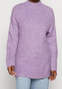 Personne portant un pull en maille douce violet clair avec poignets et ourlet côtelés, associé à un jean bleu clair déchiré, sur un fond uni.