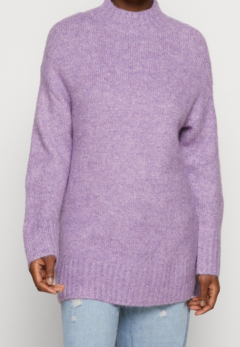 Personne portant un pull en maille douce violet clair avec poignets et ourlet côtelés, associé à un jean bleu clair déchiré, sur un fond uni.